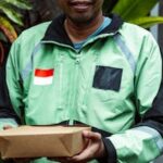 Delivery man in green jacket holding a package, smiling warmly, surrounded by lush plants.